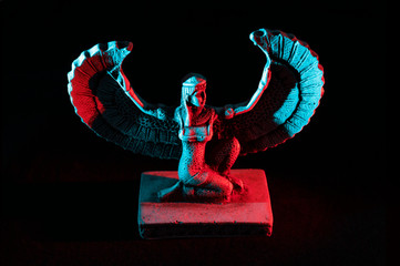 Closeup to a Isis. female old winged egyptian god mini figurine iluminated with red and blue lights over black background  - obrazy, fototapety, plakaty
