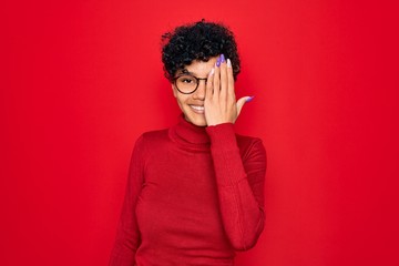 Fototapeta na wymiar Young beautiful african american afro woman wearing turtleneck sweater and glasses covering one eye with hand, confident smile on face and surprise emotion.