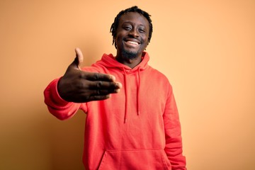 Young african american sporty man wearing sweatshirt with hoodie over yellow background smiling friendly offering handshake as greeting and welcoming. Successful business.