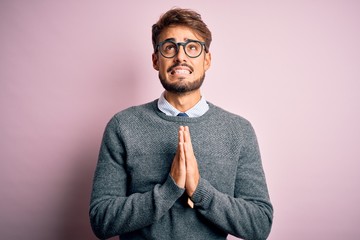 Young handsome man with beard wearing glasses and sweater standing over pink background begging and...