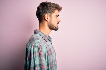 Young handsome man with beard wearing casual shirt standing over pink background looking to side,...