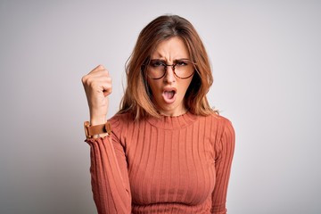 Young beautiful brunette woman wearing casual sweater and glasses over white background angry and mad raising fist frustrated and furious while shouting with anger. Rage and aggressive concept.