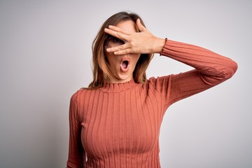 Young beautiful brunette woman wearing casual sweater and glasses over white background peeking in shock covering face and eyes with hand, looking through fingers with embarrassed expression.