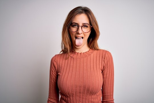 Young beautiful brunette woman wearing casual sweater and glasses over white background sticking tongue out happy with funny expression. Emotion concept.