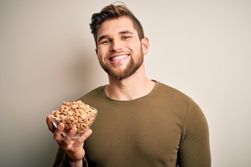 Young blond man with beard and blue eyes holding bowl with healthy salty peanuts with a happy face...
