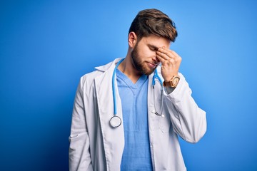 Young blond doctor man with beard and blue eyes wearing white coat and stethoscope tired rubbing nose and eyes feeling fatigue and headache. Stress and frustration concept.