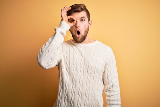 Young blond man with beard and blue eyes wearing white sweater over yellow background doing ok gesture shocked with surprised face, eye looking through fingers. Unbelieving expression.