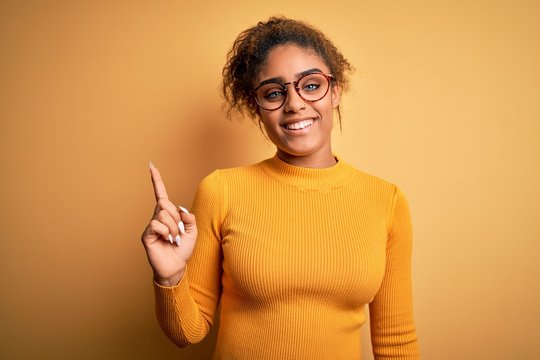 Young beautiful african american girl wearing sweater and glasses over yellow background with a big smile on face, pointing with hand finger to the side looking at the camera.