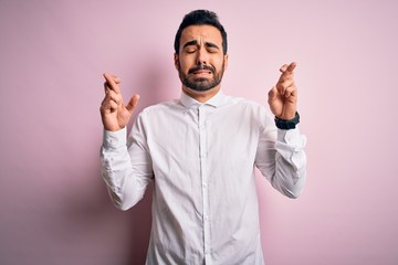 Fototapeta premium Young handsome man with beard wearing casual shirt standing over pink background gesturing finger crossed smiling with hope and eyes closed. Luck and superstitious concept.