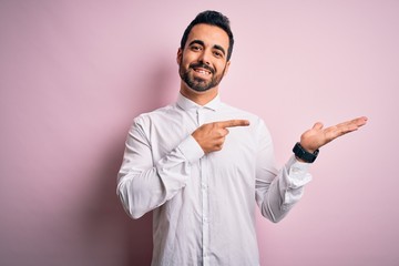 Young handsome man with beard wearing casual shirt standing over pink background amazed and smiling to the camera while presenting with hand and pointing with finger.