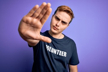 Young handsome redhead man wearing volunteer t-shirt over isolated purple background with open hand doing stop sign with serious and confident expression, defense gesture