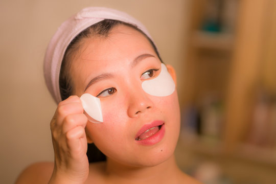 home lifestyle portrait of young happy and beautiful Asian Korean woman at bathroom mirror applying panda eye patch anti-aging product in beauty and skincare concept