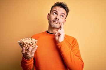 Young handsome man holding bowl with peanuts standing over isolated yellow background serious face...