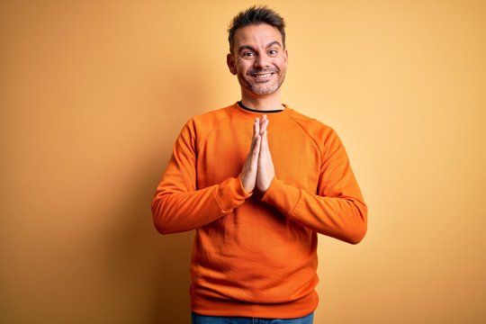Young handsome man wearing orange casual sweater standing over isolated yellow background praying with hands together asking for forgiveness smiling confident.