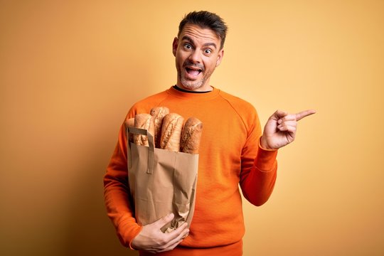 Young handsome man holding paper bag with bread over isolated yellow background very happy pointing with hand and finger to the side
