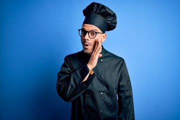 Young handsome chef man wearing cooker uniform and hat over isolated blue background hand on mouth telling secret rumor, whispering malicious talk conversation