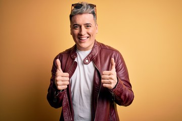 Young handsome modern man wearing fashion leather jacket and sunglasses over yellow background success sign doing positive gesture with hand, thumbs up smiling and happy. Cheerful expression