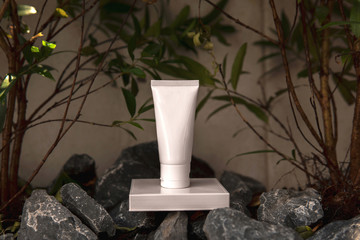 Empty treatment facial organic skincare white tube product with a blank label bottle sitting on white square pedestal in a rock garden with green leaves and natural light as a background.