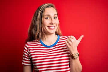 Young beautiful blonde woman wearing casual striped t-shirt over isolated red background smiling with happy face looking and pointing to the side with thumb up.