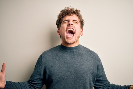 Young blond handsome man with curly hair wearing casual sweater over white background crazy and mad shouting and yelling with aggressive expression and arms raised. Frustration concept.