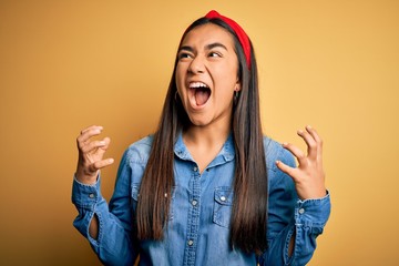Young beautiful asian woman wearing casual denim shirt and diadem over yellow background crazy and...
