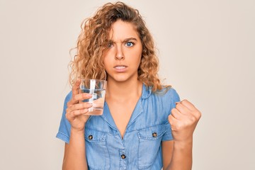 Young beautiful blonde woman with blue eyes drinking glass of water over white background annoyed and frustrated shouting with anger, crazy and yelling with raised hand, anger concept