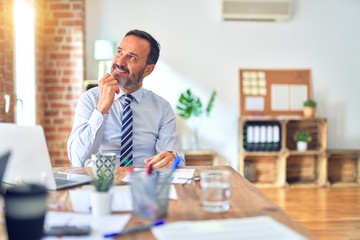 Middle age handsome businessman wearing tie sitting using laptop at the office with hand on chin thinking about question, pensive expression. Smiling with thoughtful face. Doubt concept.