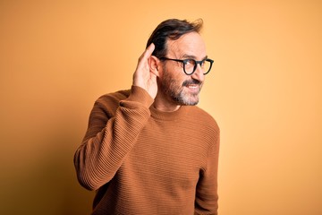 Fototapeta premium Middle age hoary man wearing brown sweater and glasses over isolated yellow background smiling with hand over ear listening an hearing to rumor or gossip. Deafness concept.