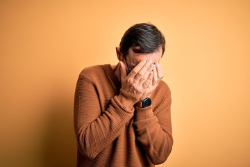 Middle age hoary man wearing brown sweater and glasses over isolated yellow background with sad expression covering face with hands while crying. Depression concept.