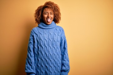 Young beautiful African American afro woman with curly hair wearing blue turtleneck sweater sticking tongue out happy with funny expression. Emotion concept.