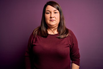 Obraz premium Beautiful brunette plus size woman wearing casual sweater over isolated purple background with serious expression on face. Simple and natural looking at the camera.