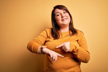 Obraz premium Beautiful brunette plus size woman wearing casual sweater over isolated yellow background In hurry pointing to watch time, impatience, upset and angry for deadline delay