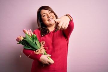 Beautiful plus size woman holding romantic bouquet of natural tulips flowers over pink background pointing fingers to camera with happy and funny face. Good energy and vibes.