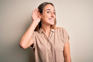 Young beautiful woman wearing casual shirt standing over isolated white background smiling with hand over ear listening an hearing to rumor or gossip. Deafness concept.