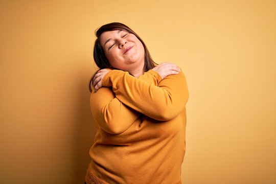 Beautiful brunette plus size woman wearing casual sweater over isolated yellow background Hugging oneself happy and positive, smiling confident. Self love and self care