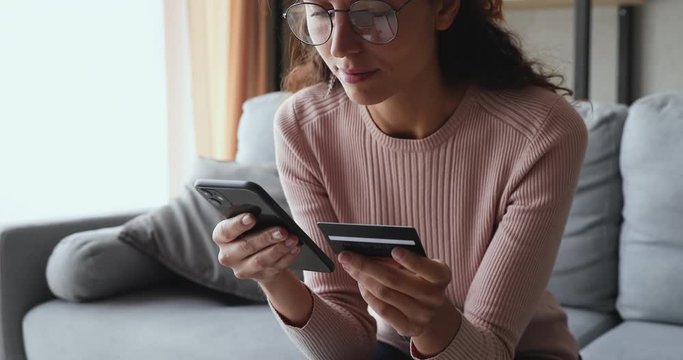 Smiling young woman customer holding credit card and smartphone sitting on couch at home. Happy female shopper using instant easy mobile payments making purchase in online store. E-banking app service