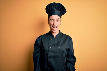 Young beautiful chef woman wearing cooker uniform and hat standing over yellow background winking looking at the camera with sexy expression, cheerful and happy face.