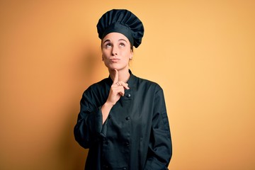 Young beautiful chef woman wearing cooker uniform and hat standing over yellow background Thinking concentrated about doubt with finger on chin and looking up wondering