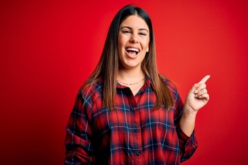 Young beautiful woman wearing casual shirt over red background with a big smile on face, pointing with hand and finger to the side looking at the camera.