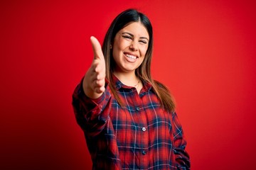 Young beautiful woman wearing casual shirt over red background smiling friendly offering handshake as greeting and welcoming. Successful business.