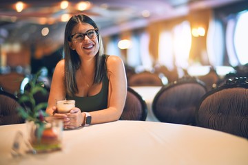 Young beautiful woman smiling happy and confident. Sitting with smile on face holding glass of coffee at restaurant