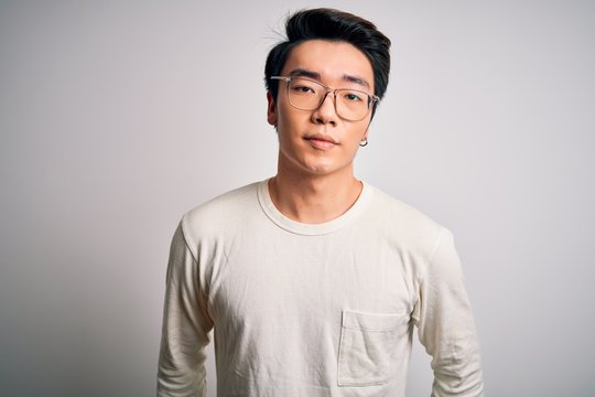 Young handsome chinese man wearing casual t-shirt and glasses over white background with serious expression on face. Simple and natural looking at the camera.
