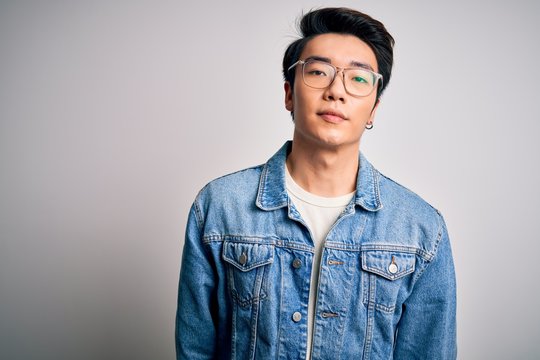 Young handsome chinese man wearing denim jacket and glasses over white background Relaxed with serious expression on face. Simple and natural looking at the camera.