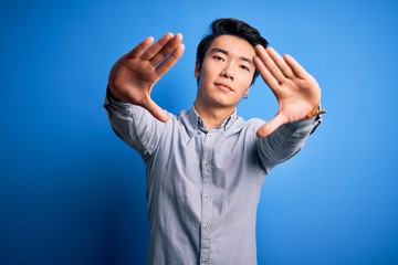 Naklejka premium Young handsome chinese man wearing casual shirt standing over isolated blue background doing frame using hands palms and fingers, camera perspective