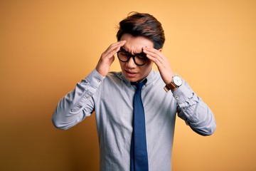 Young handsome chinese businessman wearing glasses and tie over yellow background with hand on head for pain in head because stress. Suffering migraine.