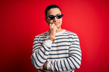 Young brazilian man wearing funny thug life sunglasses over isolated red background with hand on chin thinking about question, pensive expression. Smiling with thoughtful face. Doubt concept.