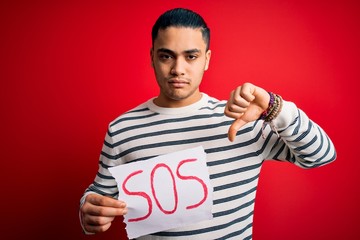 Young brazilian man with problem holding banner with sos message over red background with angry...