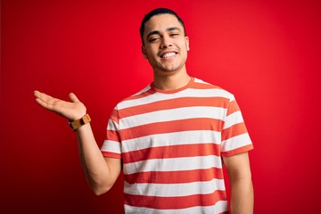 Young brazilian man wearing casual striped t-shirt standing over isolated red background smiling cheerful presenting and pointing with palm of hand looking at the camera.