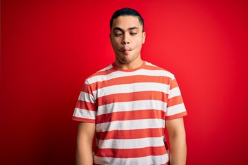 Young brazilian man wearing casual striped t-shirt standing over isolated red background making fish face with lips, crazy and comical gesture. Funny expression.