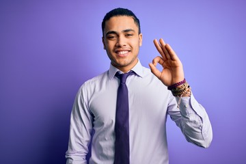 Young brazilian businessman wearing elegant tie standing over isolated purple background smiling positive doing ok sign with hand and fingers. Successful expression.